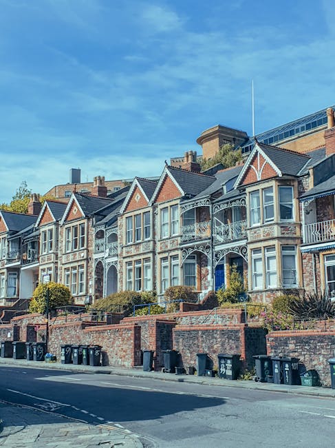 A row of Victorian-style terraced houses with ornate white metalwork balconies and bay windows, built with red brick facades and slate roofs, situated on a street with a pavement in front. The houses are elevated on a raised brick wall with landscaped garden areas featuring small bushes and trees. Several black and green wheelie bins are lined up along the roadside, with a clear blue sky overhead. The location is indicative of residential property in Dulwich, relevant to house removals and moving services provided by Dulwich Man and Van as part of a home relocation or furniture transport process.