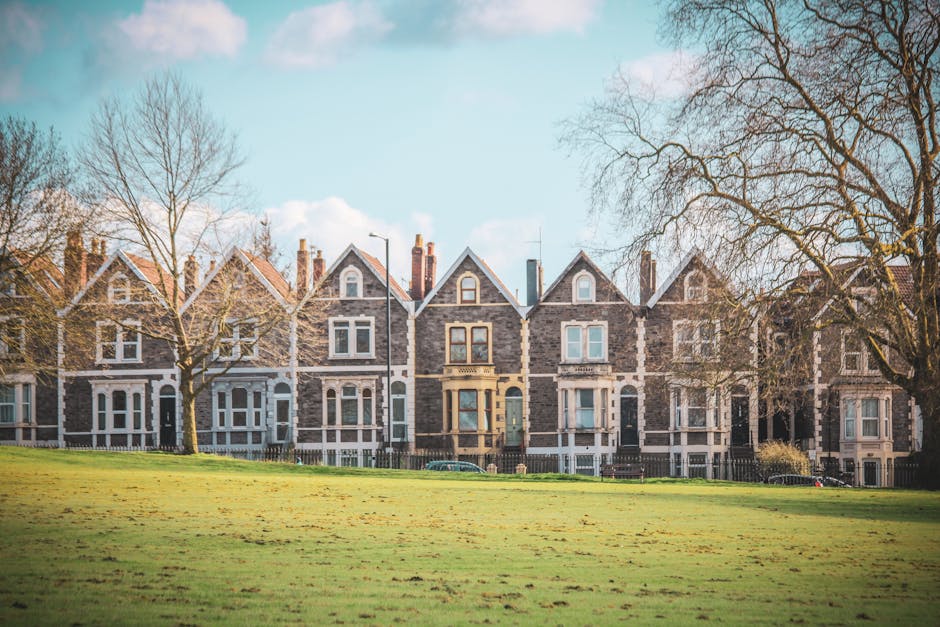 A row of Victorian-style terraced houses with stone facades, pitched roofs, and large sash windows, situated behind a well-maintained grassy area with a few leafless trees. The houses are directly accessible from the pavement, with some front gardens enclosed by low fencing, and small pathways leading to the front doors. The sky above is partly cloudy with patches of blue, providing natural daylight that illuminates the scene evenly. In the foreground, there is an open grassy area, and the scene suggests a quiet residential neighbourhood. Dulwich Man and Van's house removals services are relevant here, as these properties are typical of the type of home involved in local moving or furniture transport, often requiring careful packing, loading, and transportation logistics during a home relocation process.