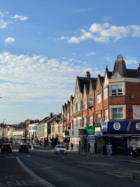A street scene in Dulwich with a row of multi-story brick houses featuring pitched roofs and bay windows, set against a blue sky with scattered white clouds. The street has moving cars and stationary vehicles at traffic lights, with pedestrians walking along the pavement. In the foreground, a Dulwich Man and Van van is parked near the curb, possibly involved in a house removal or home relocation service. Visible on the pavement are cardboard boxes, plastic wrapping, and small furniture items being prepared for transport, indicating a packing and moving process. The setting suggests an active environment where house removals, furniture transport, and local moving services are taking place, with the company occasionally supporting the loading or unloading of household belongings onto or from the vehicle during a same-day move.