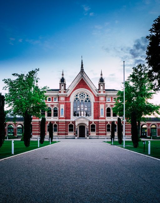 The image shows a large, historic red brick building with intricate architectural details, including arched windows, decorative stonework, and a prominent central entrance. The structure has multiple spires and a symmetrical façade, set against a blue sky with some scattered clouds. In the foreground, a wide gravel driveway leads directly to the entrance, flanked by neatly maintained green lawns and evenly spaced tall trees on either side. The scene is well-lit, capturing the building’s grandeur and the surrounding greenery. This setting illustrates a typical location that may be part of a house or building being prepared for relocation or used as a landmark in the context of moving services. Dulwich Man and Van may be involved in transporting or packing items related to the move, supporting the logistics of home relocation or furniture transport within the area.