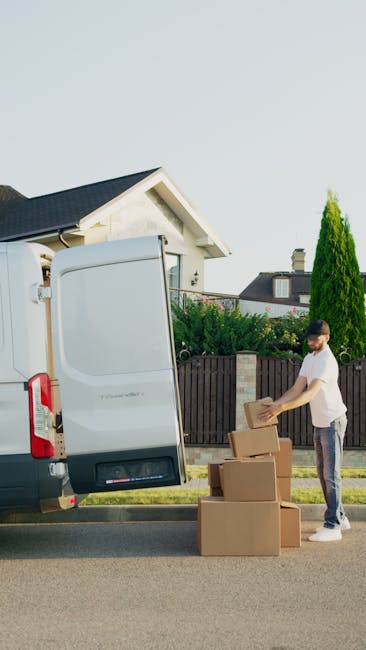 A man wearing a white t-shirt and grey pants is standing on a residential street, stacking cardboard boxes near the open rear door of a white moving van. The van is parked on the roadside, with the back doors swung open, revealing an empty cargo area prepared for the home relocation process. The boxes are of various sizes, some sealed with packing tape, and are placed on the pavement next to the van. In the background, there is a modern, single-story house with a pitched roof, white exterior walls, a small front garden with greenery, and a wooden fence separating the property from neighboring homes. The scene is well-lit with natural daylight, indicating a clear weather day, and the setting is typical of a house move involving furniture transport and packing activities. Dulwich Man and Van offers such removals services, as indicated by the scene depicting the loading process for a house move in West Dulwich.