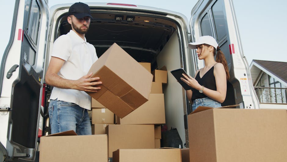 A man wearing a white polo shirt, blue jeans, and a black cap is inside the rear of a white moving van, lifting a medium-sized cardboard box. Next to him, a woman in a black sleeveless top, blue jeans, and a white baseball cap is standing beside a large cardboard box, holding a tablet device and observing the loading process. Inside the van, several cardboard boxes of various sizes are stacked on the floor, with some boxes partially visible on the pavement outside. The van has its rear doors open, revealing the interior filled with packing materials such as plastic wraps and additional boxes. The scene is set outdoors in a residential area, with a house and a clear sky visible in the background. The overall setting reflects a home relocation, with furniture and other household items being organized for transportation by Dulwich Man and Van, during the packing and loading phase of a house move at West Dulwich.