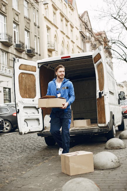 A man wearing a white t-shirt and grey pants is standing on a residential street, stacking cardboard boxes near the open rear door of a white moving van. The van is parked on the roadside, with the back doors swung open, revealing an empty cargo area prepared for the home relocation process. The boxes are of various sizes, some sealed with packing tape, and are placed on the pavement next to the van. In the background, there is a modern, single-story house with a pitched roof, white exterior walls, a small front garden with greenery, and a wooden fence separating the property from neighboring homes. The scene is well-lit with natural daylight, indicating a clear weather day, and the setting is typical of a house move involving furniture transport and packing activities. Dulwich Man and Van offers such removals services, as indicated by the scene depicting the loading process for a house move in West Dulwich.